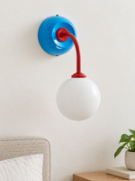 Blue + Red wall sconce with curved red arm and white glass globe, shown unlit, daylight bedside setting