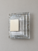 Small wall sconce with clear textured glass and white acrylic insert behind a brushed-metal cover, shown switched off in daylight